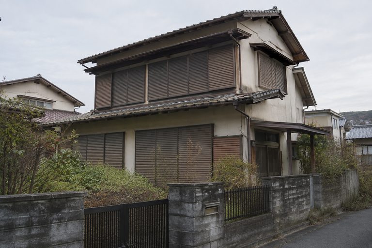 雨戸が閉まったままの古い木造2階建ての住宅とブロック塀